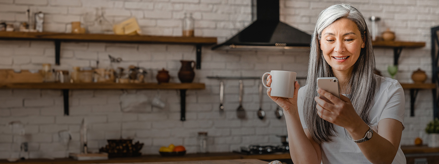 woman in kitchen looking holding mug looking at cell phone