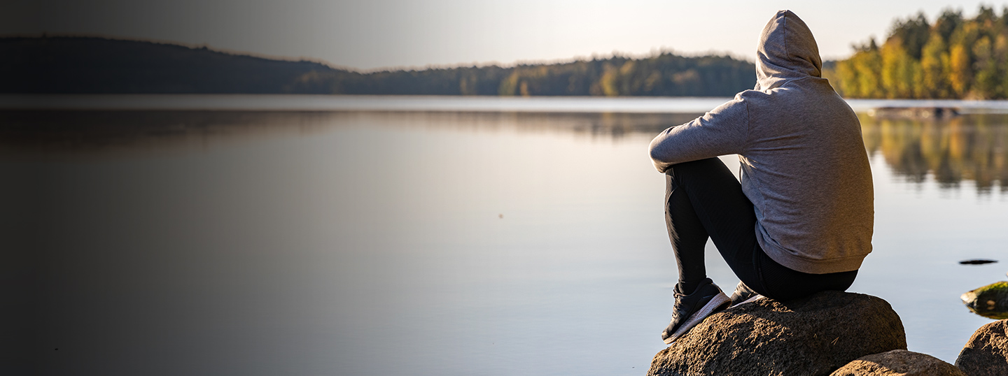 person in hoodie sitting on rock by near a lake